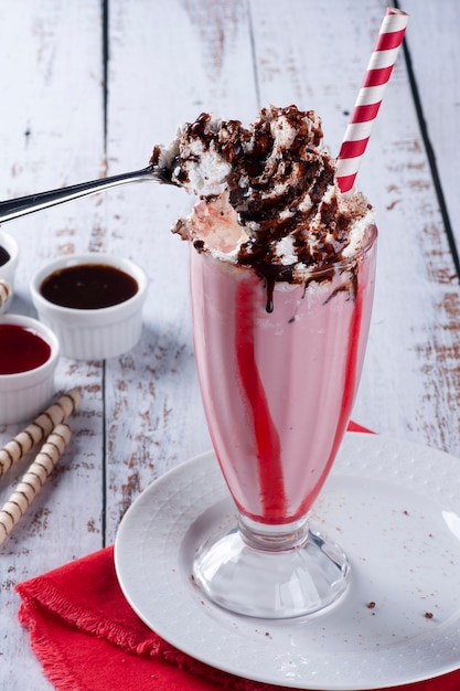 Beautiful glass of strawberry milkshake with whipped cream and\
chocolate sauce. a spoon taking the whipped cream