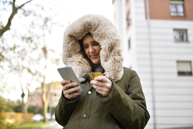 Photo beautiful and good looking middle aged woman in winter shopping with credit card and smart phone in the city