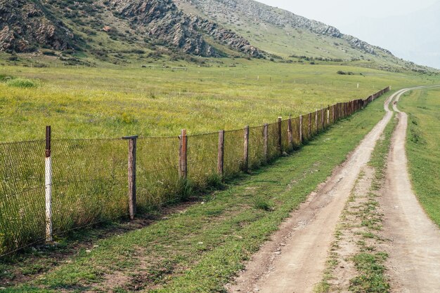 Beautiful green mountain landscape with long dirt road along fence and big mountains in fog. Atmospheric foggy mountain scenery with dirt road among rocks and big mountains. Length road in countryside