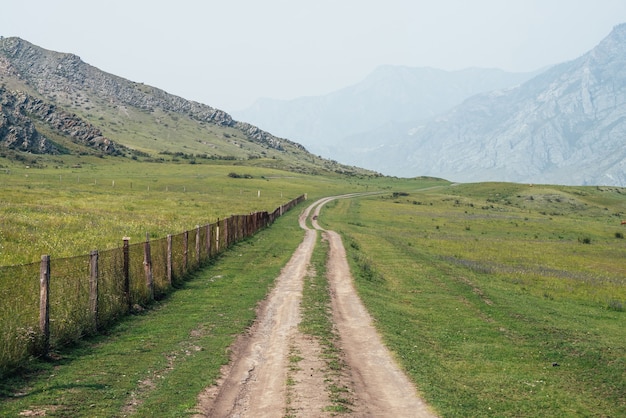 Beautiful green mountain landscape with long dirt road along fence and big mountains in fog. Atmospheric foggy mountain scenery with dirt road among rocks and big mountains. Length road in countryside