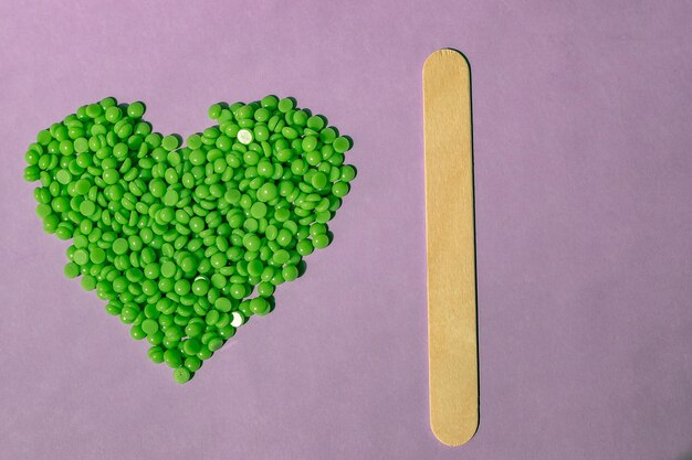 Beautiful green wax granules with Aloe extract for depilation are poured on a light purple background in the shape of a heart wooden spatula Epilation depilation removal of unwanted hair