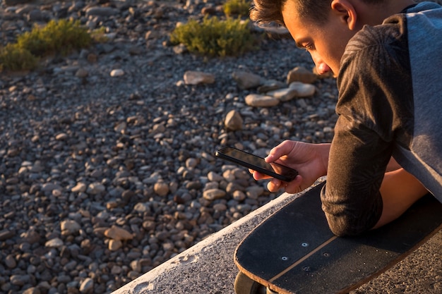 Foto bel ragazzo e adolescente in spiaggia da solo con il suo telefono - giocando, chattando o guardando video con il suo skateboard