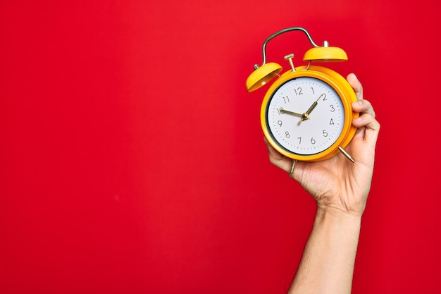 Beautiful hand of man holding vintage alarm clock over isolated red background