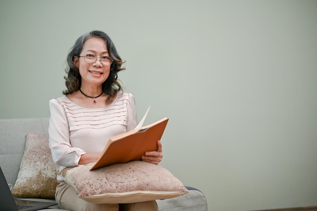 아름답고 행복한 60대 은퇴한 아시아 여성이 안경을 쓰고 거실에서 책을 읽고 있다