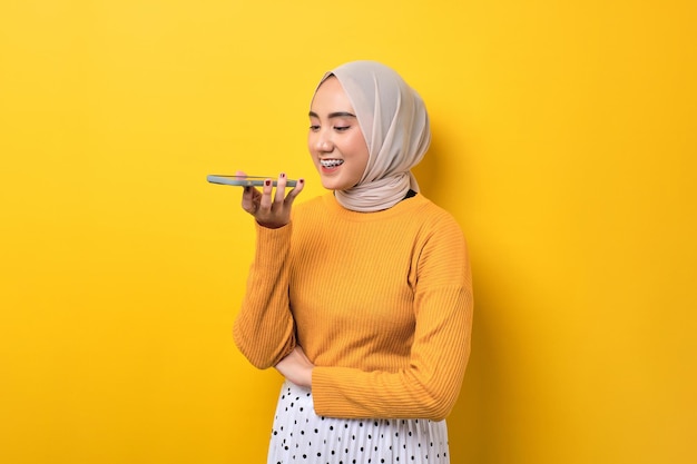 Beautiful happy Asian girl wearing hijab talking on mobile phone in loudspeaker mode isolated on yellow background