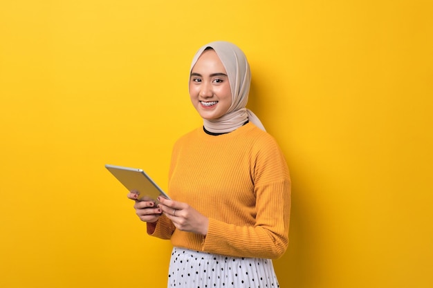 Beautiful happy Asian girl wearing hijab using digital tablet and smiling at camera isolated on yellow background