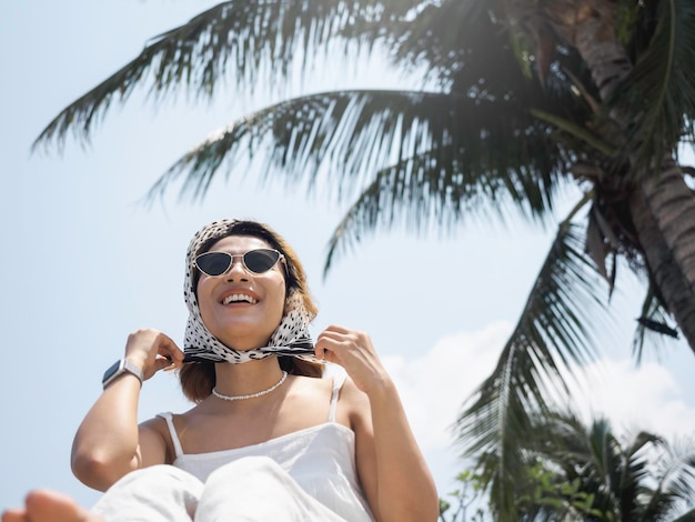 Красивый счастливый азиатский портрет женщины в повседневной белой рубашке в солнцезащитных очках и шарфе для волос наслаждается улыбкой на пляже под кокосовыми пальмами и фоном голубого неба летом
