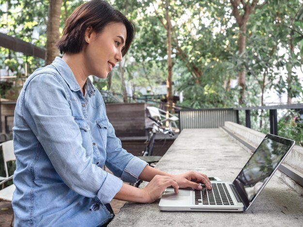 Beautiful happy asian woman short hair wearing blue jeans shirt
using laptop computer in cafe with green background