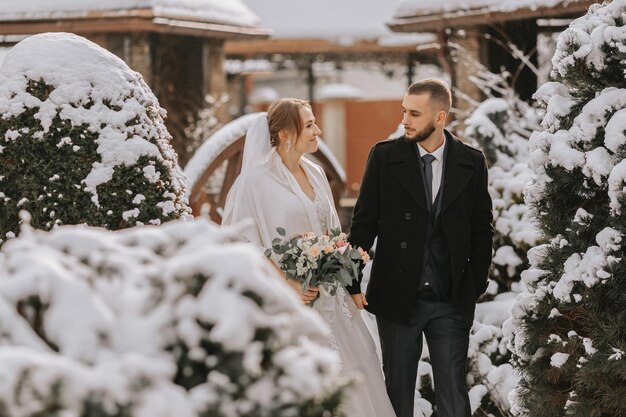 Foto la splendida e felice sposa e sposo camminano tra gli alberi coperti di neve la sposa e lo sposo nel parco invernale la sposa con un bouquet di fiori in un vestito da sposa e un poncho lo sposo in un cappotto nero