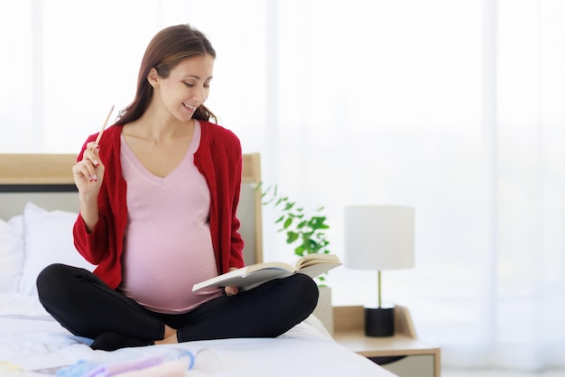 Una bella madre caucasica felice è seduta sul suo letto con in mano un taccuino che tiene un elenco di tutte le forniture e i vestiti in preparazione per la nascita del neonato