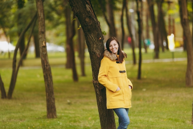 Beautiful happy caucasian young smiling brown-hair woman in yellow coat, jeans, boots in green forest. Fashion female model with fall golden leaves standing and walking in early autumn park outdoors.
