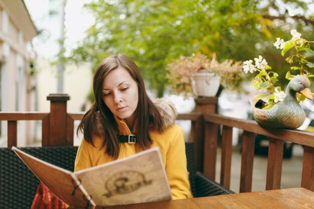 Bella felice giovane caucasica sorridente donna dai capelli castani in cappotto giallo leggendo, ordinando dal menu in un ristorante all'aperto o in un caffè vicino alla strada e decidendo cosa mangiare a cena nella stagione autunnale.