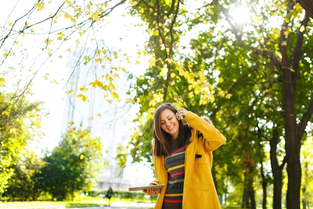 The beautiful happy cheerful brown-haired woman in a yellow coat and striped longsleeve rejoices with a tablet in her hands and white headphones in fall city park on a warm day. Autumn golden leaves.