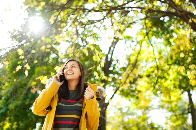 La bella donna dai capelli castani allegra felice in un cappotto giallo e maniche lunghe a righe parlando al cellulare nel parco cittadino autunnale in una giornata calda. foglie d'oro autunnali.