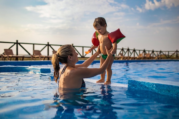 La bella mamma allegra e felice gioca con il suo piccolo figlio carino e divertente con maniche colorate luminose in una piscina blu intenso con acqua limpida e trasparente, in estate