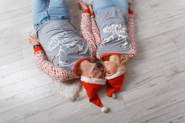 Beautiful happy couple in Christmas scenery. Weared in funny sweaters and hats. Laying down on the floor and kissing.