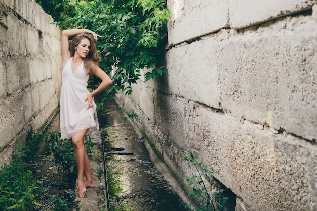 Bella ragazza di dancing felice con capelli naturali ricci in vestito bianco vicino alle foglie verdi dell'albero. ritratto di bellezza estiva. la donna dei sogni ama la natura. donna ispirata con vista da sogno nel lungo tunnel nella foresta