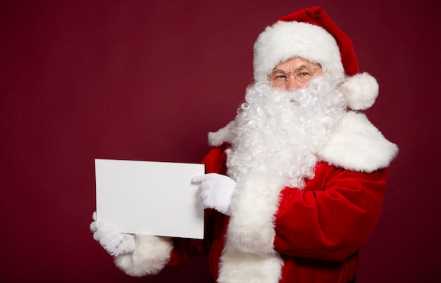 Beautiful happy excited Santa Claus is posing and showing white blank board in hands 