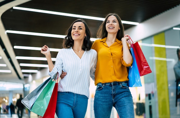 Le belle amiche giovani felici ed emozionate con i sacchetti di carta stanno camminando intorno al centro commerciale