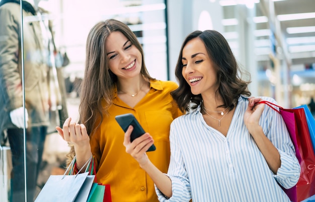 Belle amiche giovani felici ed emozionate con sacchetti di carta e smart phone stanno camminando per il centro commerciale