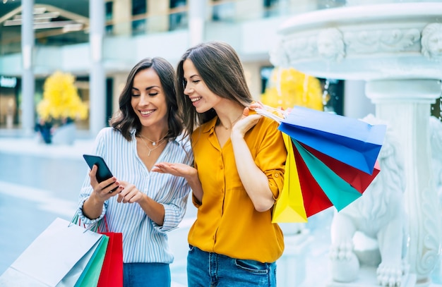 Beautiful happy and excited young girl friends with paper bags and smart phone are walking around the shopping mall