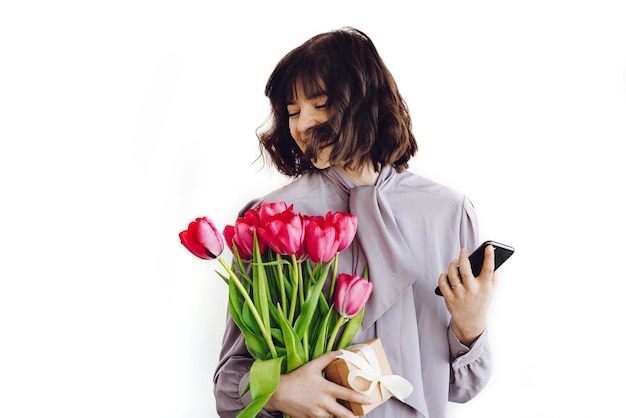 Photo beautiful happy girl having fun and smiling holding red tulips and phone on white background indoors space for text stylish young brunette woman laughing and waving hair happy womens day
