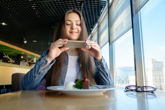 美しい幸せな女の子は、カフェで食べ物、テーブルの上のラテ、デザートアイスクリームチョコレートケーキチェリーミントの写真を作っています