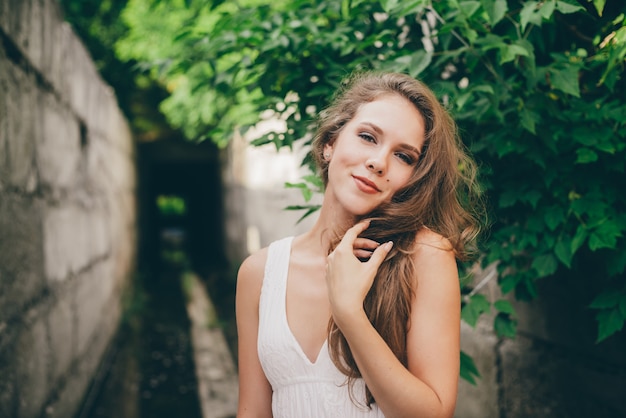 Beautiful happy girl with curly natural hair in white dress near green tree leaves