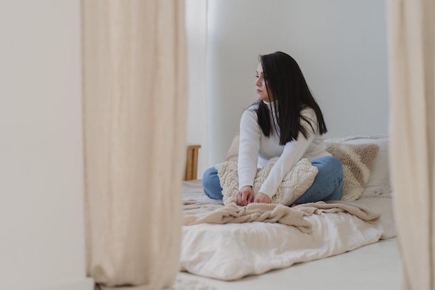 긴 머리를 가진 아름다운 행복한 소녀는 파스텔 색상의 인테리어로 베개를 껴안고 침대에 앉아 있습니다