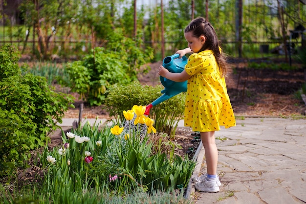 Photo a beautiful happy girl in a yellow dress is watering yellow flowers from a watering can in the garde...