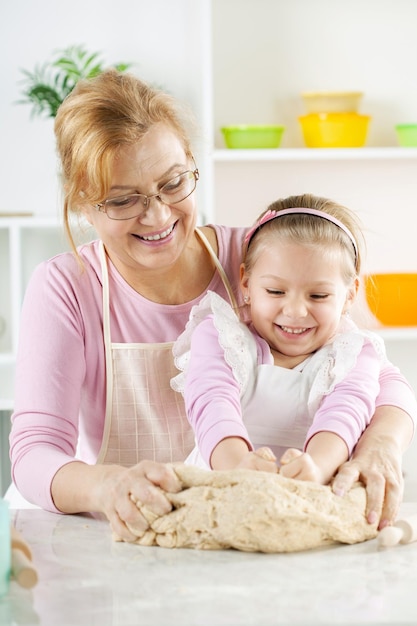 Красивая, счастливая бабушка и внучка вместе месят тесто на кухне.