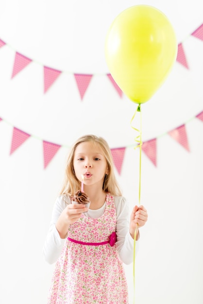 Beautiful happy little girl holding balloon and small birthday cake with candle. Looking at camera.