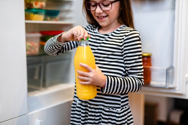 Beautiful and happy little girl taking bottle of fresh orange juice from refrigerator.