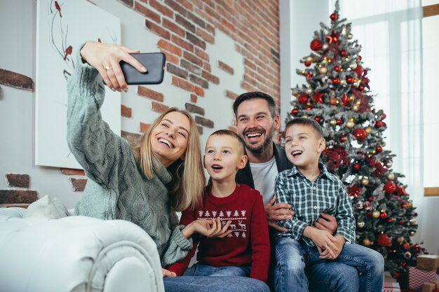 Beautiful happy married couple with two children using smart phone talking to friends or family congratulates Merry Christmas by video call conference app. Modern technology New Year 2022 congrats.