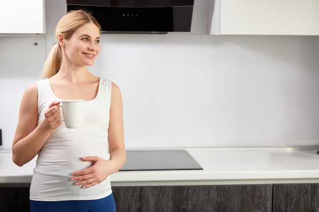 Beautiful happy pregnant woman drinking cup of tea or coffee in kitchen at home in the morning