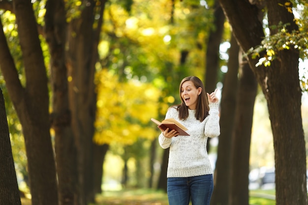 The beautiful happy smiling brown-haired woman in white sweater standing with a red book in fall city park on a warm day. Autumn golden leaves. Reading concept.