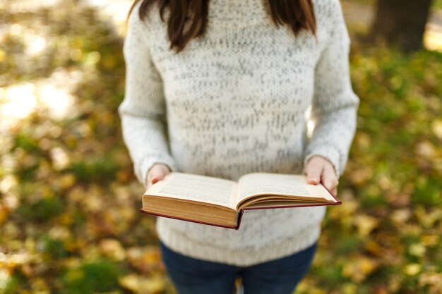 The beautiful happy smiling brown-haired woman in white sweater standing with a red book in fall city park on a warm day. Autumn golden leaves. Reading concept.