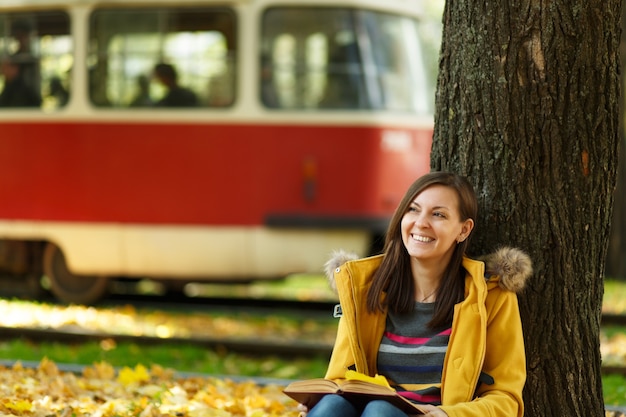 Foto la bella donna dai capelli castani sorridente felice in cappotto giallo e jeans seduta sotto l'albero di acero con un libro rosso nel parco cittadino autunnale. sfondo del tram. foglie d'oro autunnali. concetto di lettura