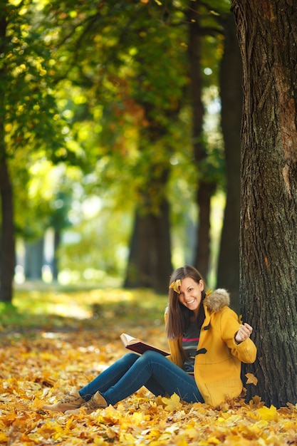 The beautiful happy smiling brown-haired woman in yellow coat and jeans sitting under the maple tree with a red book in fall city park on a warm day. Autumn golden leaves. Reading concept