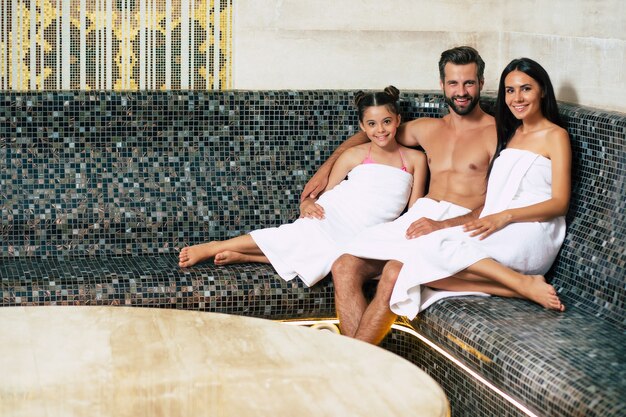 Beautiful happy smiling family in bath towels are looking on camera while relaxing in sauna or hammam. Vacation and Spa procedure