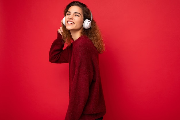 赤の上に分離された濃い赤のセーターを着て美しい幸せな笑顔の若いブルネットの巻き毛の女性