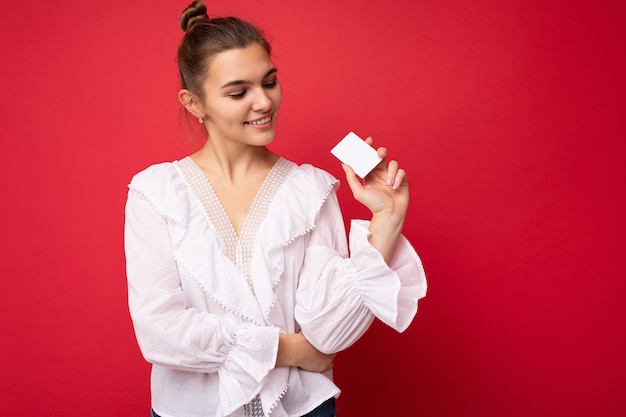 Bella felice sorridente giovane donna bionda scura che indossa camicetta bianca isolate su sfondo rosso in possesso di carta di credito guardando la carta di plastica. copia spazio