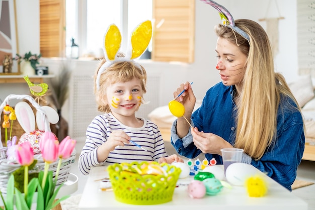 Foto bellissimo figlio felice e madre in fasce con orecchie da coniglio decorano le uova con un pennello e vernici luminose si preparano a celebrare la pasqua si divertono a casa