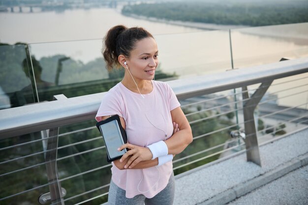 Beautiful happy sporty girl with earphones standing with crossed arms on the city bridge and smiling toothy smile looking away. Athlete woman resting after intense cardio workout outdoors