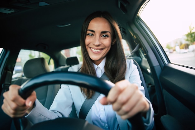 Beautiful happy successful businesswoman is driving a new modern car in good mood. Portrait cute female driver steering car with safety belt