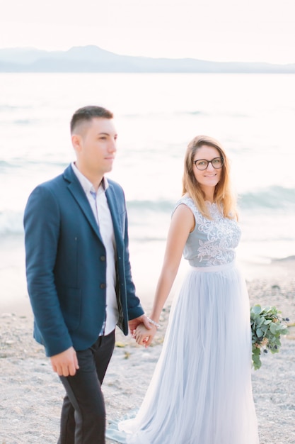 Beautiful happy wedding couple of groom in blue jacket and bride in luxury blue dress walking holding hands near the Garda lake. Italy, Sirmione