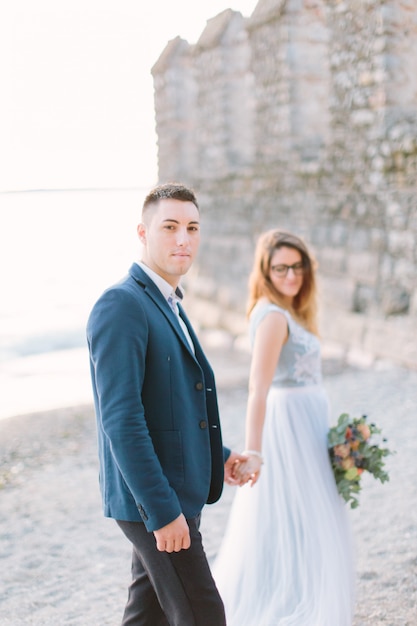Beautiful happy wedding couple of groom in blue jacket and bride in luxury blue dress walking holding hands near the Garda lake. Italy, Sirmione