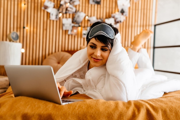 Beautiful happy woman in bed using laptop over white blanket