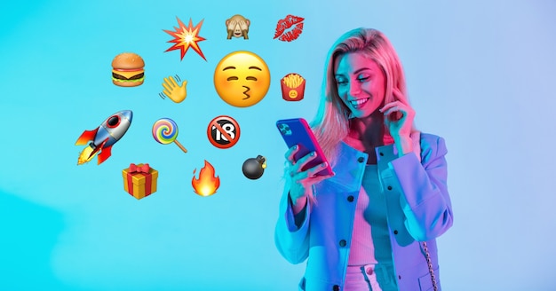Photo beautiful happy woman holding smartphone with flat emoji on neon background. social media emoji communication concept