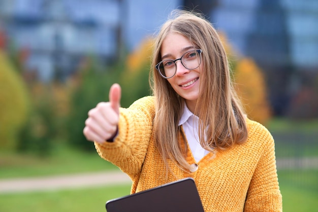 美しい幸せな女性、前向きな若い大学生または大学生のガラスのティーンエイジャーの女の子が公園に座っており、大学のキャンパスはジェスチャーのように親指を立てて屋外で笑っています。教育